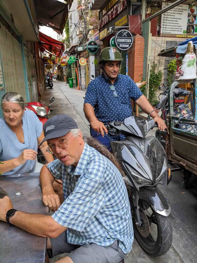 two people sitting at a table with a scooter passing close by